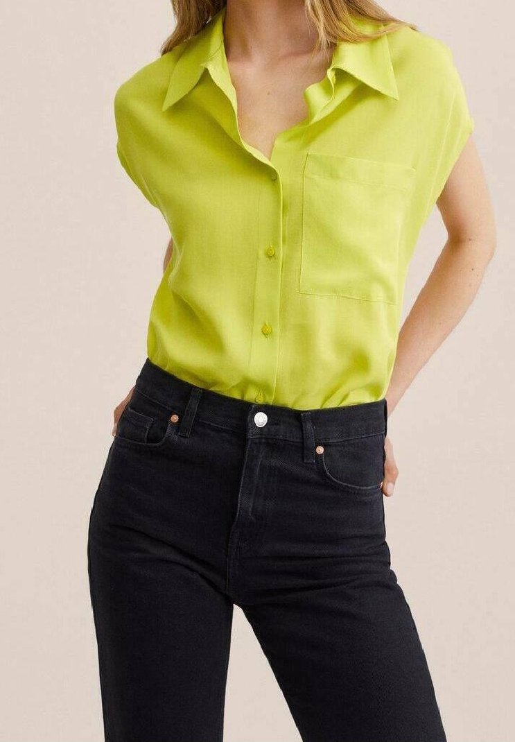 Blusa a maniche corte di un vivace verde lime con chiusura a bottoni e una singola tasca sul petto, abbinata a jeans neri a vita alta.