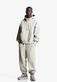 Light gray hoodie with a front pocket and drawstring, paired with matching gray sweatpants. Model wears gray sneakers with red accents.