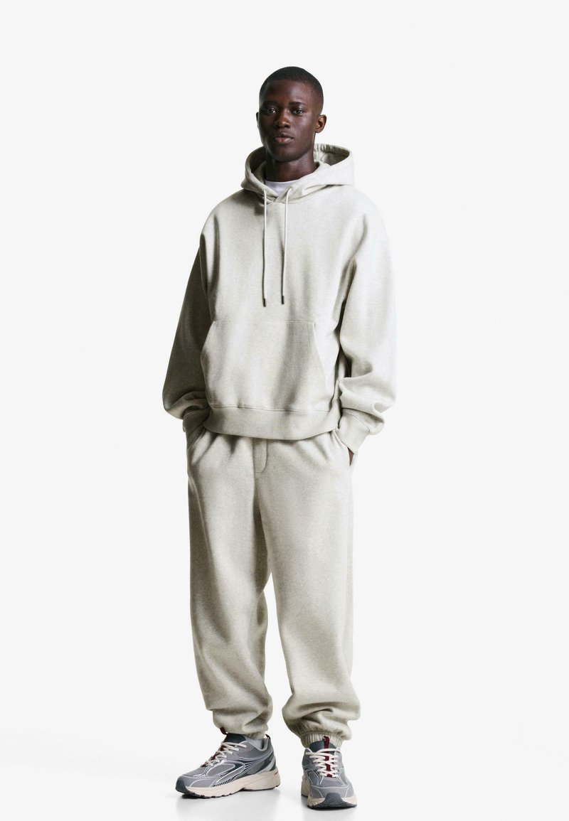 Light gray hoodie with a front pocket and drawstring, paired with matching gray sweatpants. Model wears gray sneakers with red accents.