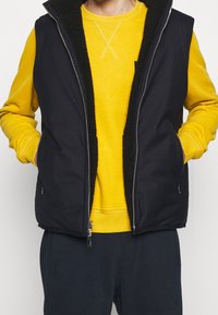 Svart ärmlös väst med ett plyschsvart foder, med dragkedja framtill och sidofickor, bärs över en gul tröja.