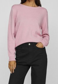 Kvinna i en ljusrosa stickad tröja med ribbade ärmar och svarta högmidjade byxor, med en hand i fickan, mot en enfärgad bakgrund.