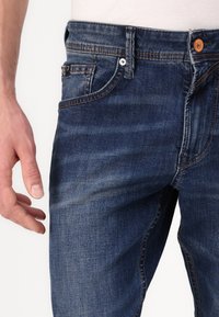 Jeans in denim blu scuro con una texture sfumata, design a cinque tasche, chiusura con bottone di rame e dettagli di cucitura a contrasto.