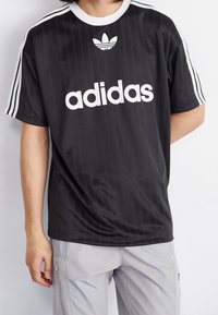 Černé tričko Adidas s bílým logem a třemi pruhy na rukávech, má kulatý výstřih a jemnou vertikální texturu. Kombinováno se šedými kalhotami.
