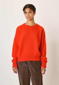 Jeune femme aux cheveux tressés portant un pull oversize rouge vif et un pantalon en velours côtelé marron, debout devant un fond clair uni.