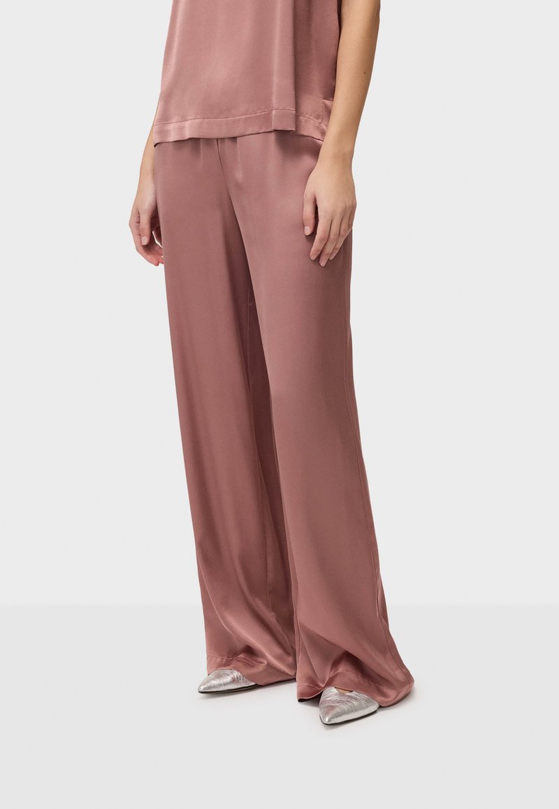 Personne portant un pantalon large et fluide en satin rose poussiére avec un haut assorti et des chaussures argentées à bout pointu, debout devant un fond clair uni.