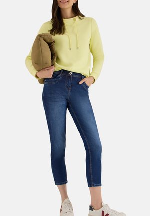 Kvinde iført gul hættetrøje og korte blå jeans, holder en brun jakke, med den ene hånd i lommen og sneakers på hvid baggrund.