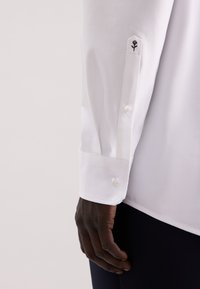 Camicia bianca a maniche lunghe con polsini abbottonati, caratterizzata da un piccolo ricamo di una rosa nera sulla manica. Tessuto di cotone liscio, vestibilità aderente.