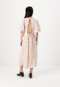 Crème jurk met een bloemenpatroon, korte puffmouwen en een grote uitsnede op de rug, taillering met een strikdetail. Draagt zwart lederen hakken.