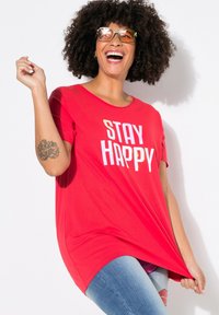 Rode t-shirt met korte mouwen met de tekst "STAY HAPPY" in het wit, gecombineerd met blauwe jeans. De stof lijkt zacht, met een relaxte pasvorm en ronde hals.