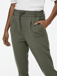 Γυναίκα που φοράει παντελόνι ελιάς με κορδόνι και πλαϊνές τσέπες, και μια λευκή υφασμάτινη κοντομάνικη μπλούζα, με το χέρι στην τσέπη, όρθια μπροστά σε λευκό φόντο.