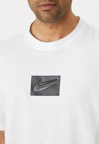 Bílá bavlněná tričko s texturovanou černou koženou nášivkou s vyraženým logem Nike na hrudi. Kulatý výstřih a krátké rukávy.