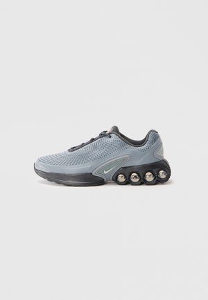 Chaussure de sport grise avec un dessus en maille texturée, semelle noire et accents métalliques. Présente un design de rembourrage unique avec des éléments arrondis.