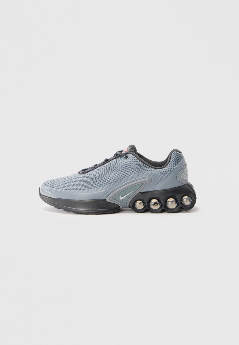 Chaussure de sport grise avec un dessus en maille texturée, semelle noire et accents métalliques. Présente un design de rembourrage unique avec des éléments arrondis.