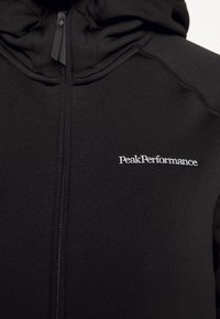 Sudadera con cremallera negra y cuello alto, hecha de una tela suave. Presenta un logo bordado de "PeakPerformance" en el pecho.