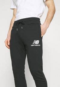 Černé tepláky z bavlněné směsi, s úzkým pasem na šňůrku a bočními kapsami. Logo New Balance je vytištěno bíle na levém stehně.