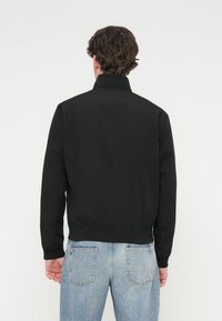 Μαύρο μπουφάν με γιακά που στέκεται, υφή υφάσματος, ριμπ μανσέτες και τελείωμα, και καθαρό σχέδιο στην πλάτη. Συνδυασμένο με τζιν σε ανοιχτό γαλάζιο χρώμα.