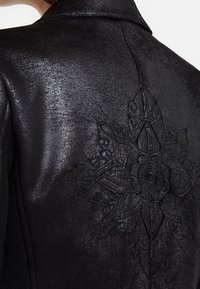 Veste en cuir noir avec une texture brillante et une broderie florale noire complexe dans le dos, mettant en avant le souci du détail et l'artisanat.