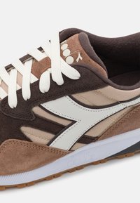 Bruna och beiga mockaskor med vita snören, vit sidremsa och vadderad krage, med den präglade "diadora"-logotypen på den vita sulan.