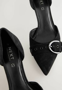 Mules noirs en daim à bout pointu avec une large sangle ornée de œillets en métal et d'un accent en boucle argentée. Doublure intérieure lisse en noir.