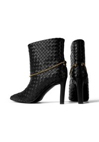 Czarne botki na obcasie z cholewką do kostki, wykonane z plecionej skóry, ozdobione złotym łańcuchem wokół kostki. Szpiczasty nosek, smukła sylwetka.