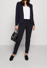 Navy blazer and matching joggers with white side stripes, paired with a white top and a black crocodile-pattern handbag. Black loafers.