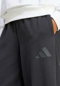 Des sweatpants en coton gris foncé présentent une taille élastique, une poche latérale et un logo Adidas brillant et ton sur ton sur la jambe gauche.