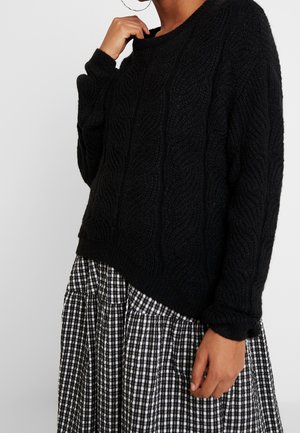 Personne portant un pull noir en tricot torsadé et une jupe plissée à carreaux noirs et blancs, tenant le col du pull d'une main.