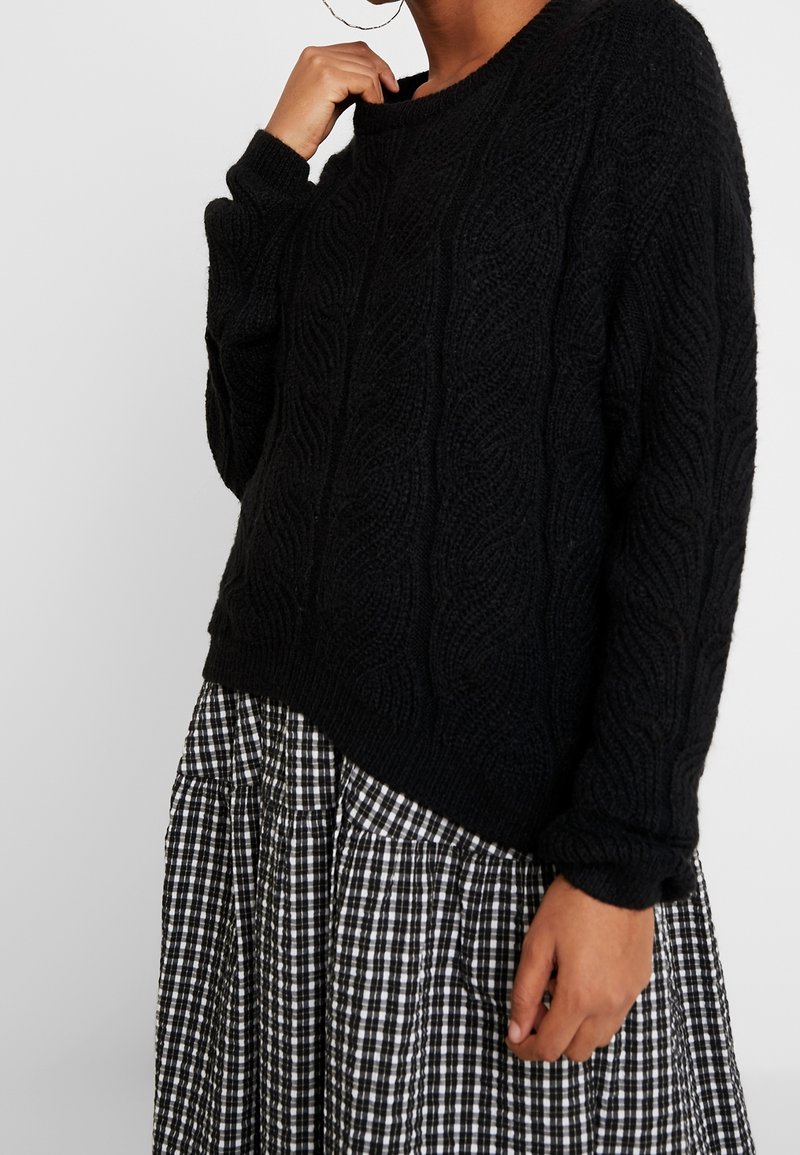 Personne portant un pull noir en tricot torsadé et une jupe plissée à carreaux noirs et blancs, tenant le col du pull d'une main.