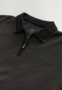Polo negro con diseño de media cremallera, cuello texturizado y un sutil patrón geométrico en tonos marrón oscuro y claro. Etiqueta de la marca visible.