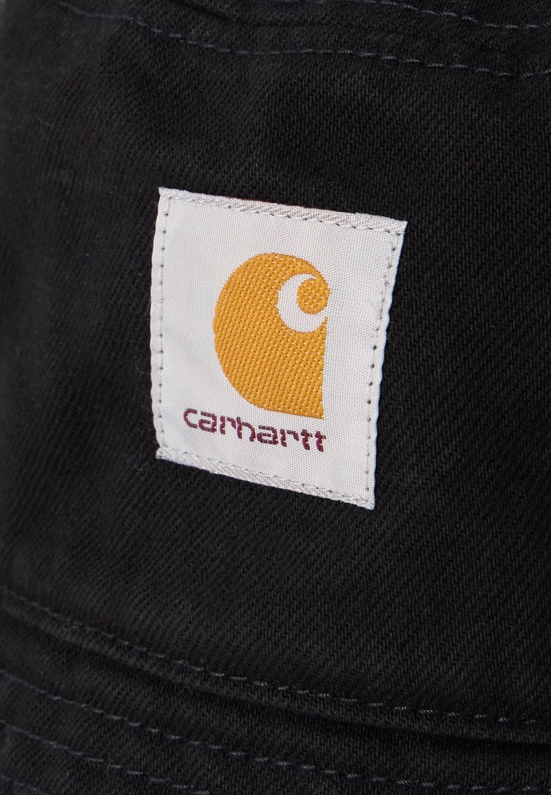 Quadratisches weißes Stoffetikett mit einem gelben stilisierten "C" und dem Text "carhartt", auf schwarzen strukturierten Stoff genäht.