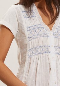 Donna che indossa una blusa bianca con texture e motivi geometrici ricamati in blu, con maniche corte arrotolate e bottoni sul davanti.