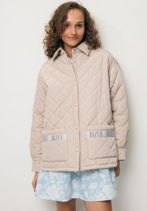 Vrouw draagt een beige doorgestikte jas met voorzakken met het label "BIZUU" over een blauwe bloemenrok, staand tegen een effen achtergrond.