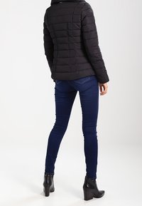 Svart vadderad jacka med horisontellt quiltat mönster, för ett par mörkblå smala jeans. Modellen bär svarta ankelskorn.