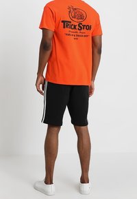T-shirt en coton orange avec un texte graphique noir et un design de camion au dos. Associée à des shorts noirs avec des rayures blanches sur les côtés. Baskets blanches.