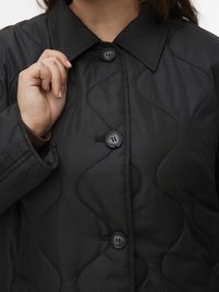 Γυναίκα που φοράει μαύρο καπιτονέ μπουφάν με μεγάλα κουμπιά, κρατώντας το γιακά με το αριστερό της χέρι κοντά στον λαιμό της.