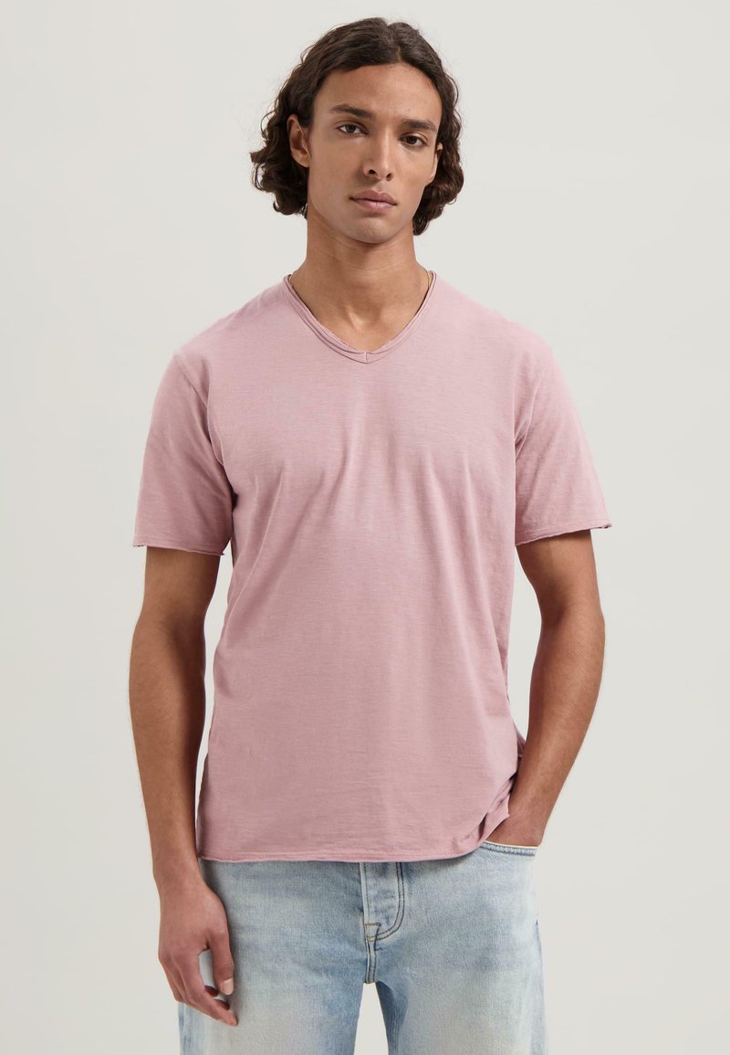 Camiseta rosa de manga corta con escote en V, hecha de una tela suave. El modelo está de pie con las manos en los bolsillos, vistiendo jeans de mezclilla de color azul claro.