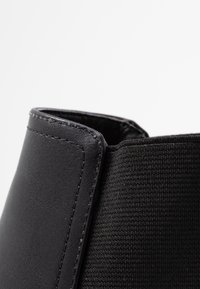 Czarny botek za kostkę z gładką skórzaną cholewką i teksturowanym paneliem z tkaniny. Przeszycia wzdłuż szwu dodają kontrastu.