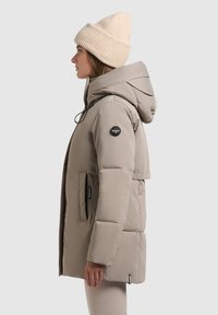 Padded, zip-up jakna u svijetloj bež boji s kapuljačom, s crnim zakrpun na rukavu i bočnim džepovima. Rebrasta bež kapa na glavi.