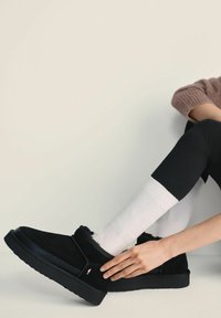 Czarne zamszowe buty typu slip-on z gumową podeszwą, zdobione przeszywanym wzorem i małym złotym akcentem, w zestawie z białymi skarpetkami żebrowanymi.