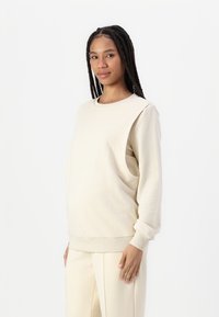 Sudadera beige de manga larga, con escote redondo y un diseño de corte único en un hombro. Textura suave y corte holgado.