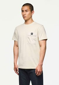 Mężczyzna w beżowym t-shircie z krótkim rękawem i kieszonką na klatce piersiowej, w ciemnych dżinsach, stojący na tle jednolitego białego tła.