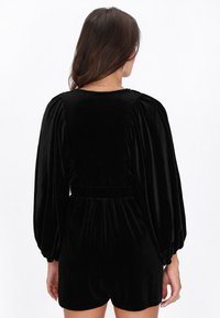 Fekete bársony overall, puffos ujjakkal, derékban húzott szabással és lágy tapintású anyaggal. A fazon egyszínű nyakkivágással és rövid szárakkal rendelkezik.