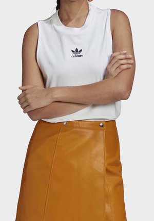 Vrouw die een witte mouwloze Adidas top draagt met een zwart logo en een hooggetailleerde mosterdgele leren rok met metalen knoopdetails.