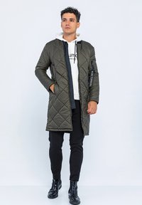 Gequilteter olivgrüner langer Mantel mit einem Frontreißverschluss, ausgestattet mit seitlichen Taschen und einem kontrastierenden schwarzen Reißverschluss. Trägt man über einem weißen Hoodie und schwarzen Hosen.