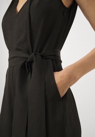 Svart jumpsuit med V-hals, bredt stoffbelte knyttet i livet, og sidelommer med en glatt, teksturert stofffinish.