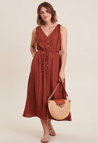 Eisenrotes, ärmelloses Midi-Kleid mit V-Ausschnitt, vorderen Knöpfen und einem verstellbaren Taillengürtel. Kombiniert mit einer Strohtasche und flachen Sandalen.