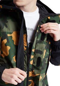 Kamouflagejacka i gröna och bruna nyanser, dragkedjeslutning och kardborreankare. Fodrad insida med en tryckt vit skjorta under.