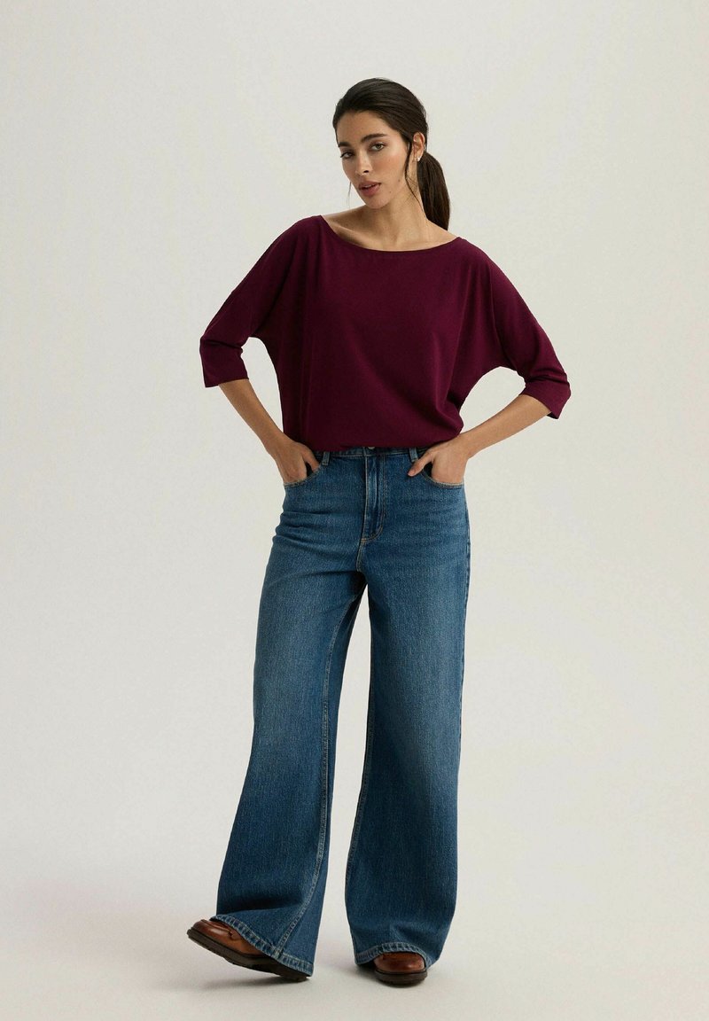 Burgundowa, oversize'owa bluzka z szerokim dekoltem w łódkę, w połączeniu z niebieskimi dżinsami o wysokim stanie i rozszerzonym fasonie. Całość dopełniają brązowe buty.