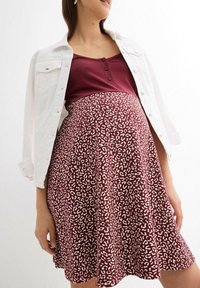 Robe de maternité bordeaux avec un imprimé léopard blanc ; longueur genoux, forme A, assortie à une veste en denim blanche et un top bordeaux en dessous.