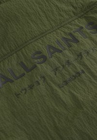 Alyvų žalia medžiaga su tekstūruota paviršiumi ir tinklelio raštu. Išraiškingai iškilios juodo spalvos raidės skelbia "ALLSAINTS" ir "LONDON."